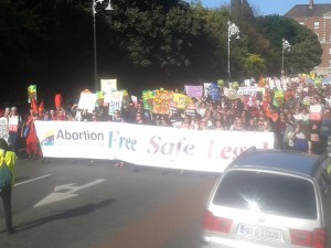 Ag teacht isteach ar thaobh dheis Chearnóg Mhuirfeann (tabhair faoi deara an placárd i nGaeilge sa lár taobh láimhe deise)